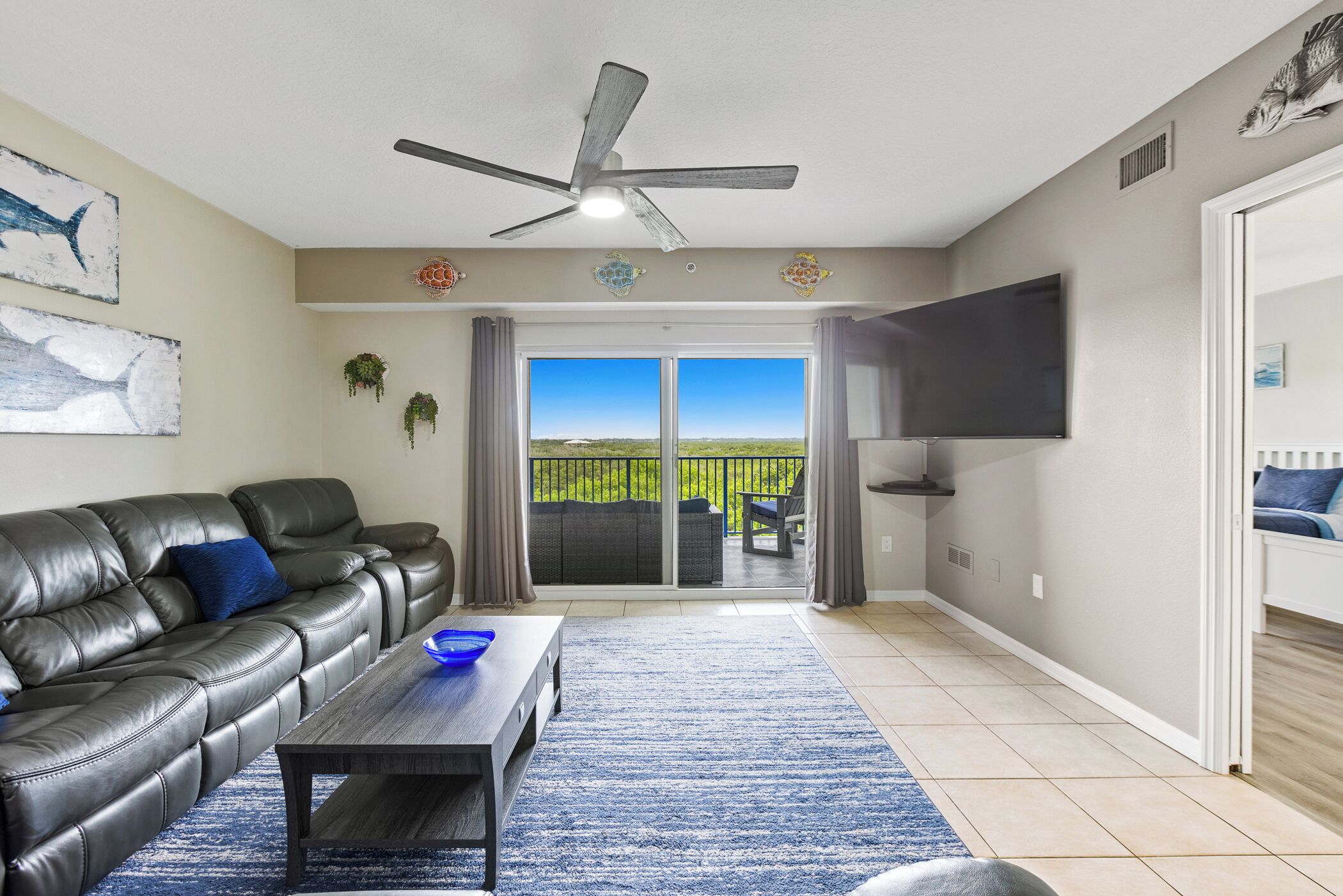 A living room with a leather sofa, a coffee table, a ceiling fan, a TV, and a view through a sliding glass door to a patio.