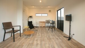 Un vaste espace de vie doté d’un plancher en bois, d’une chaise noire, d’une table en bois, d’une petite chaise jaune, d’un téléviseur posé sur un meuble et d’une fenêtre.