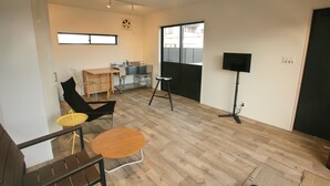 Une pièce avec un plancher en bois, un bureau, une chaise, un tabouret, un haut-parleur noir et une fenêtre.