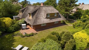 A thatched-roof house with a brick extension, surrounded by lush greenery and a well-maintained lawn. - Avenarius Hüs - Reet. Kampen. Garten (Kampen (Sylt))