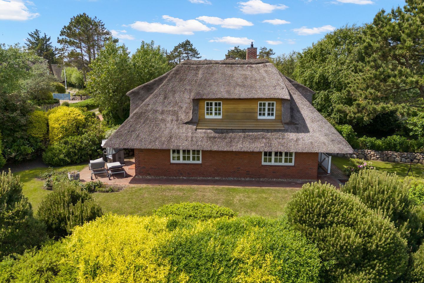 A thatched-roof house with a brick exterior, surrounded by lush greenery and trees.