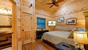 Ein Zimmer einer Holzhütte mit einem Bett, einem Fernseher, einem Badezimmer mit Waschbecken und einem Deckenventilator.