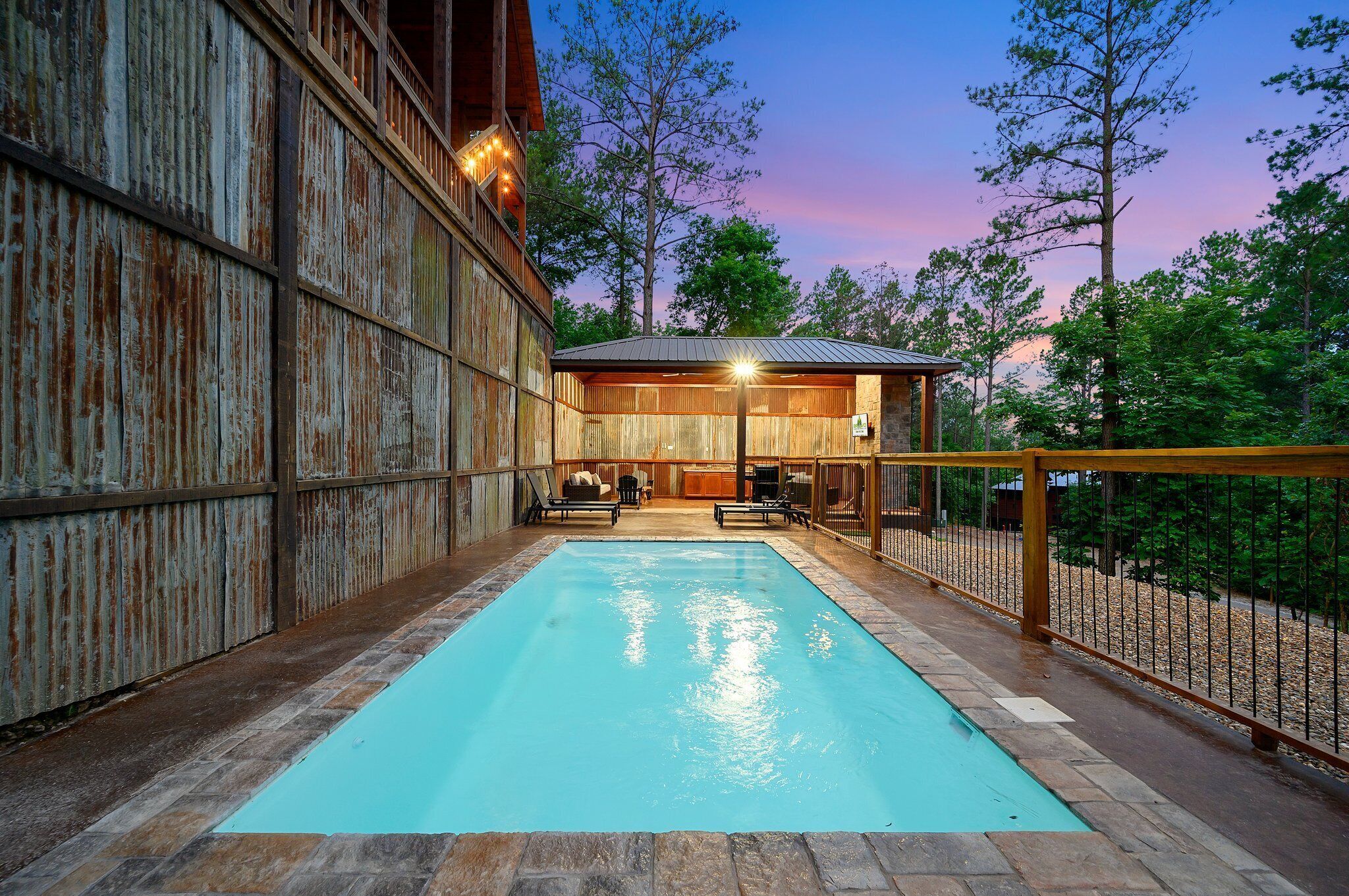 A swimming pool with a wooden deck, a covered seating area, and a rustic building with a metal exterior.