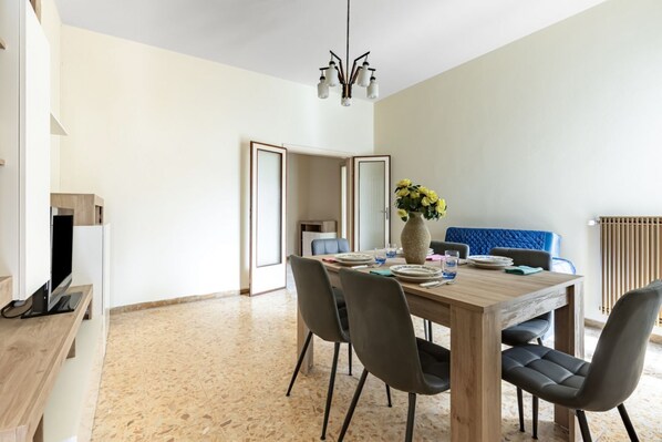 A dining area with a wooden table, chairs, a vase with flowers, and a television on a stand.