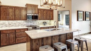 A modern kitchen with wooden cabinets, a stainless steel stove, a large island with stools, and a hanging light fixture. - Custom home close to ski resort with hot tub (Angel Fire)