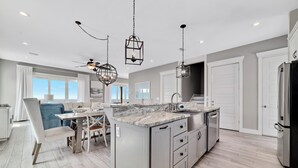 A modern kitchen with a central island, white cabinetry, and a dining area with a blue chair. - "Sandy Feet Retreat" | Beachfront | Private Pool | Beach Getaways (Pensacola)