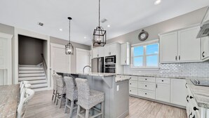 A modern kitchen with white cabinets, a central island with bar stools, a built-in oven, and a window overlooking the outside. - "Sandy Feet Retreat" | Beachfront | Private Pool | Beach Getaways (Pensacola)