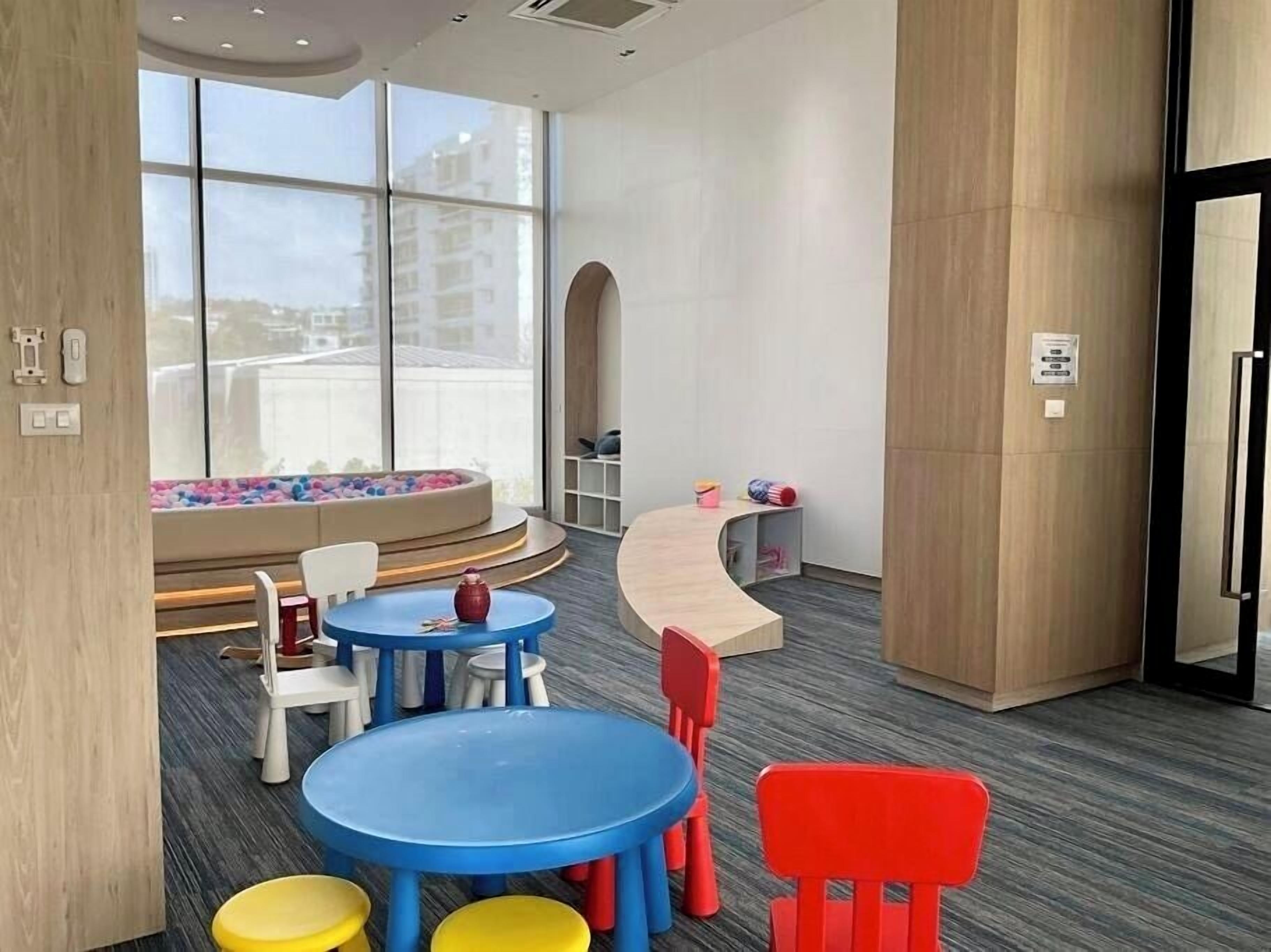 A modern hotel lobby with a curved reception desk, colorful children's play area, and large windows offering a view of the city.