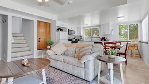 A modern living room with a sofa, coffee table, and dining area. There is a staircase leading to another level, a kitchen with white cabinets, and a large window providing natural light. - Oasis Condo on the fairway; fast WiFi, smart TVs, spectacular golf course views (Victorville)