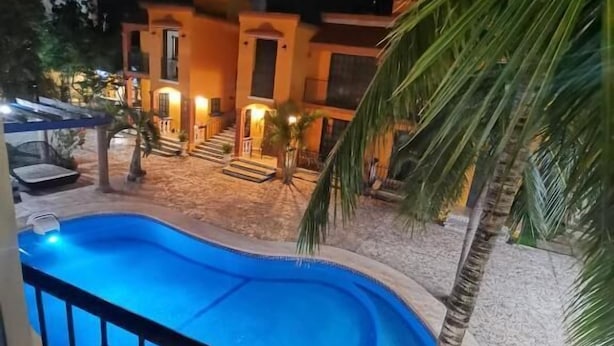 Ein Swimmingpool mit einer Steinterrasse, umgeben von Palmen und einem Gebäude mit warmer Beleuchtung.