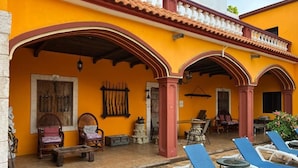 Ein gelbes Gebäude mit einer überdachten Terrasse, ausgestattet mit Holzmöbeln, einem Tisch und blauen Lounge-Stühlen.