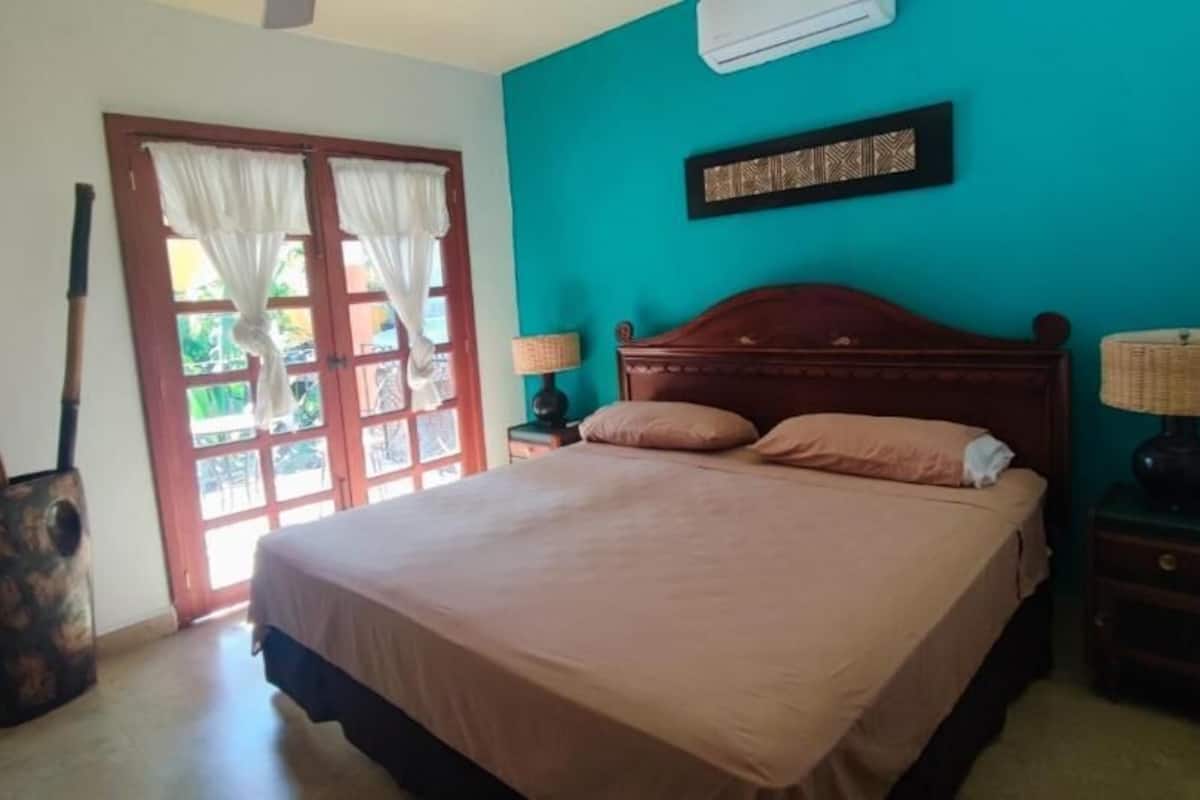 Un dormitorio con cama, mesita de noche, lámpara, silla de madera y ventana con cortinas.
