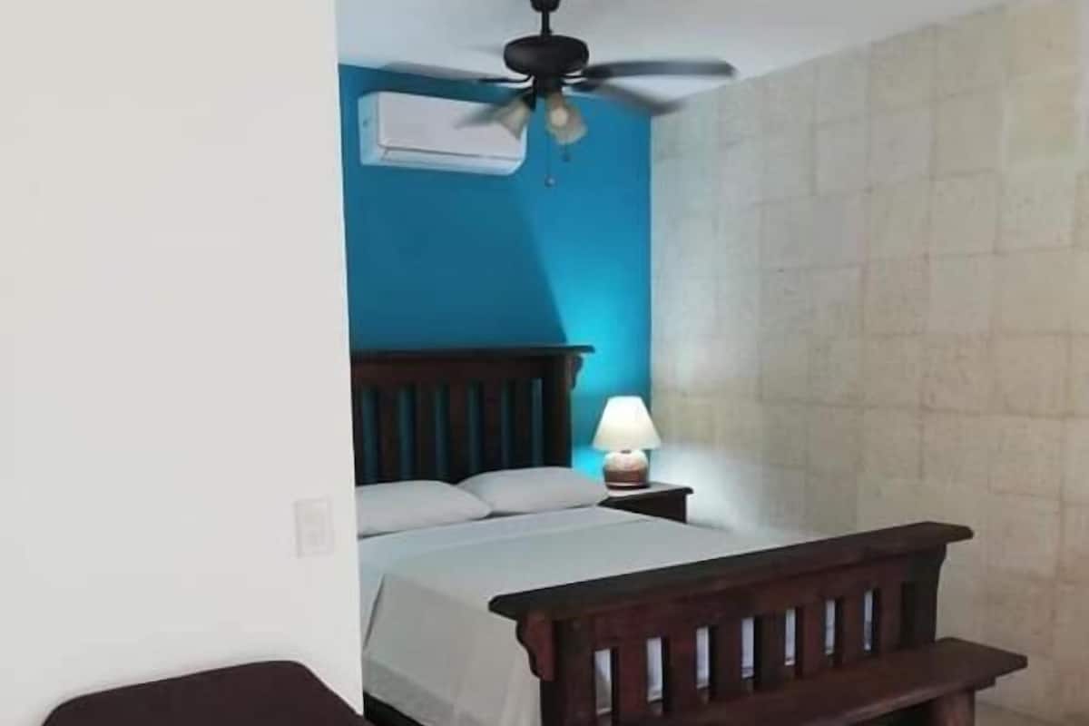Un dormitorio con una cama, un banco de madera, una silla, una mesita de noche con lámpara y un ventilador de techo.