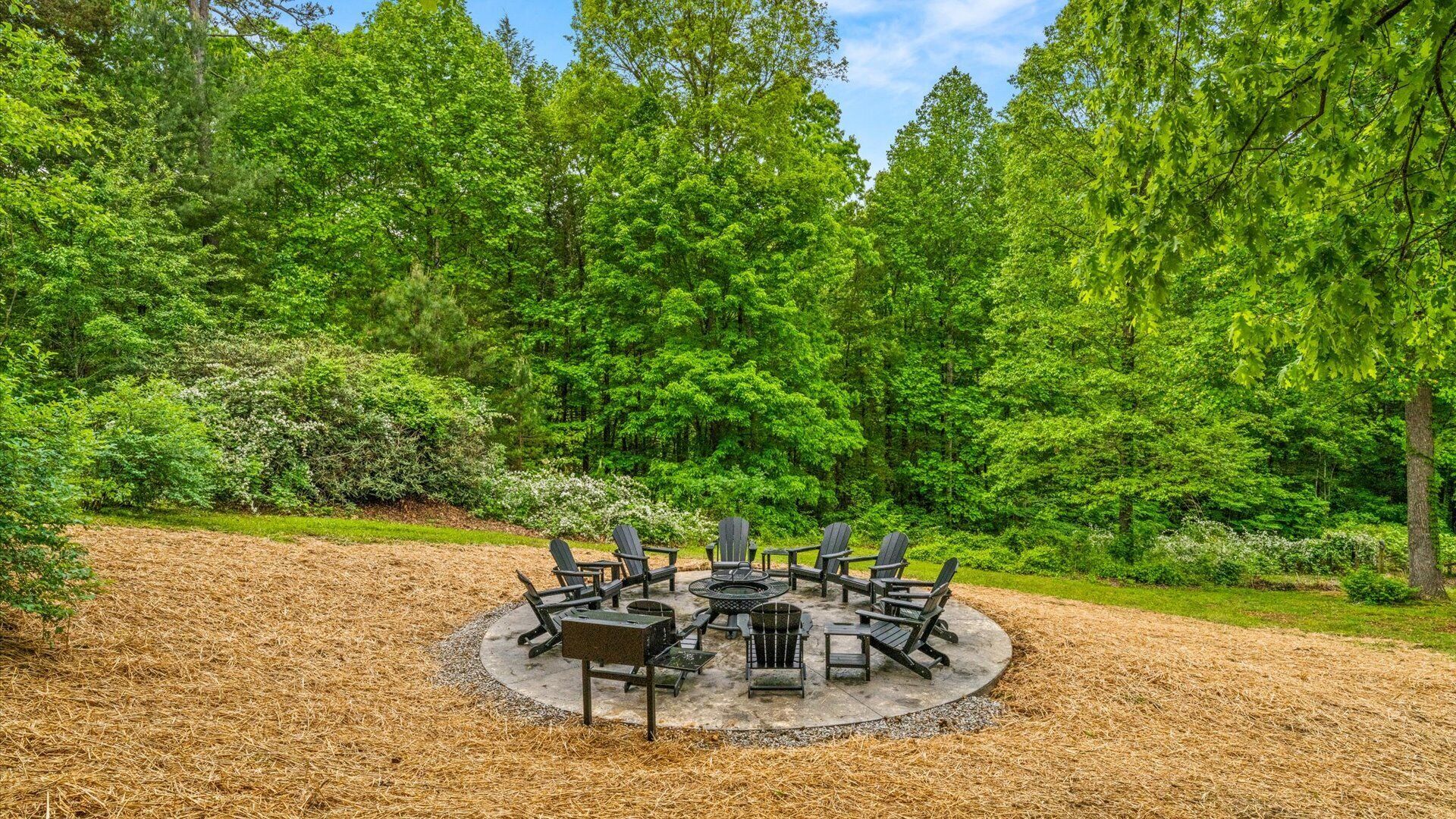 A circular outdoor seating area with Adirondack chairs and a fire pit, surrounded by a mulched ground and dense green trees.