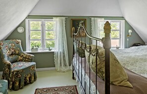 Un dormitorio con cama con dosel, un sillón floral y vistas a la vegetación por las ventanas.