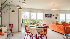 A modern dining area with a round table, red chairs, and a glass-top table. There is a large window with a view of greenery and a white ceiling with a unique light fixture. - Les Pieds dans l'eau by Interhome (Cléder)