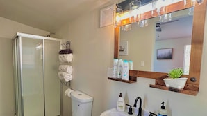 A bathroom with a glass shower enclosure, a toilet, a sink with a mirror above it, and various toiletries on the wall. - Cozy and Quiet Tiny House Cottage with Skylight 2 Minutes from Main Street (Julian)