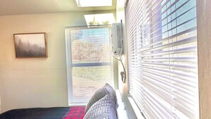 A bedroom with a skylight, a bed with a red and black quilt, a window with blinds, and a wall-mounted thermostat. - Cozy and Quiet Tiny House Cottage with Skylight 2 Minutes from Main Street (Julian)