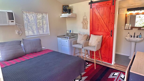 A bedroom with a bed, a desk, a chair, a red door, a ceiling fan, and a window with blinds. - Cozy and Quiet Tiny House Cottage with Skylight 2 Minutes from Main Street (Julian)