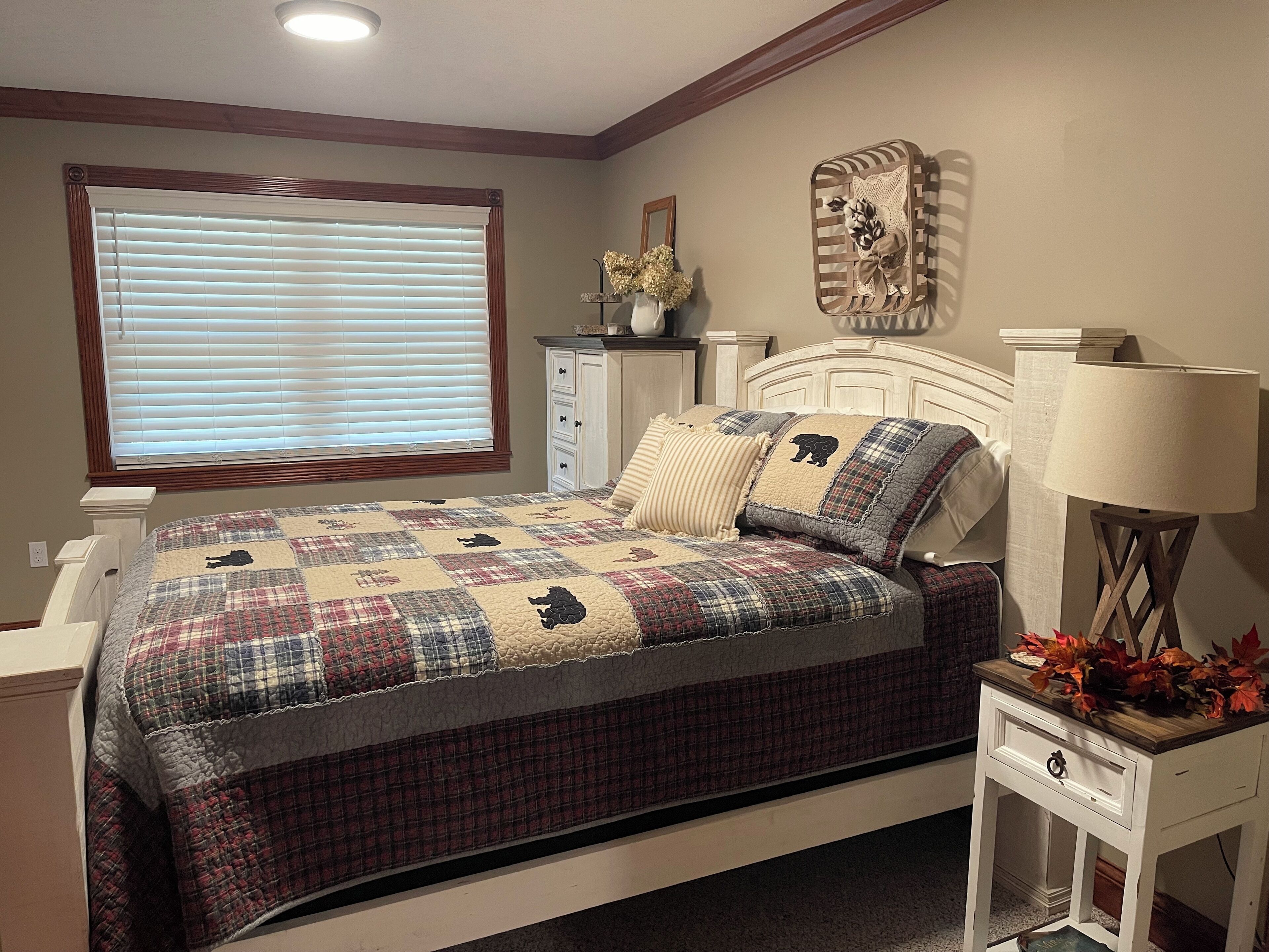 A bedroom with a bed, a nightstand, a lamp, a dresser, a window with blinds, and a decorative wall piece.