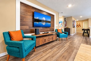 A modern living room with a flat-screen TV mounted on a wooden wall, two teal armchairs, a wooden coffee table, and a pool table.