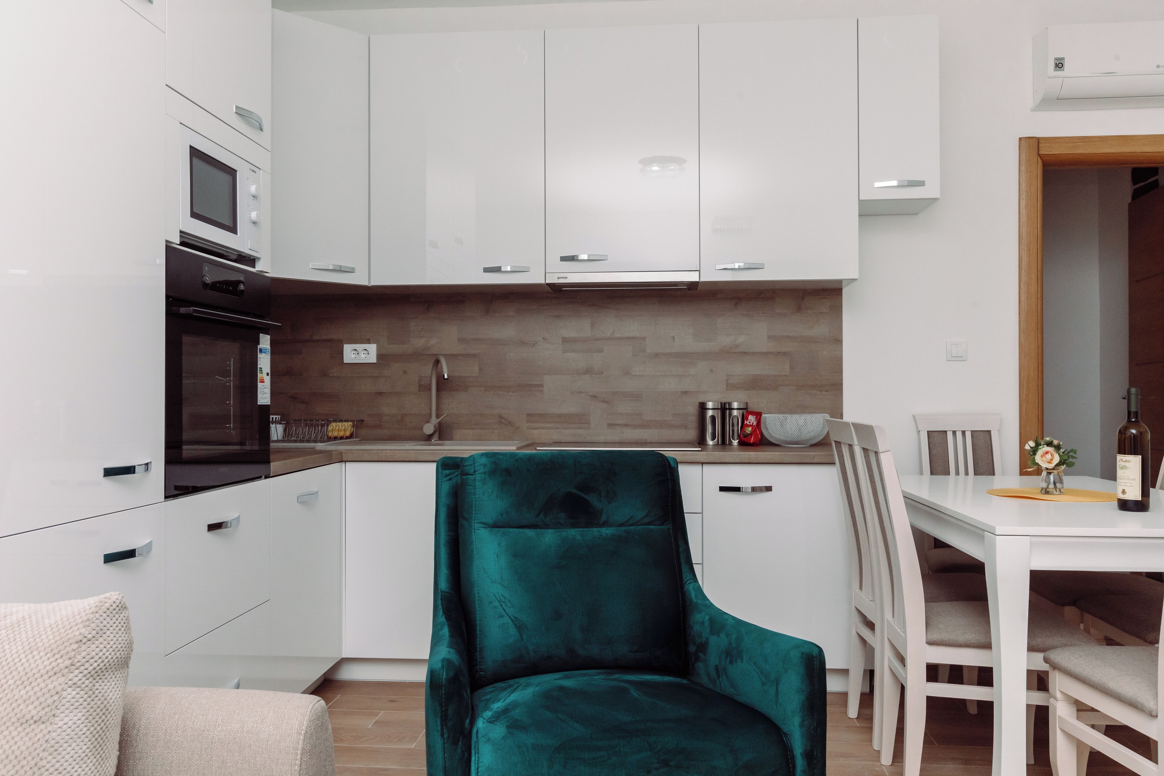 A modern kitchen with white cabinets, a built-in oven, and a teal chair.