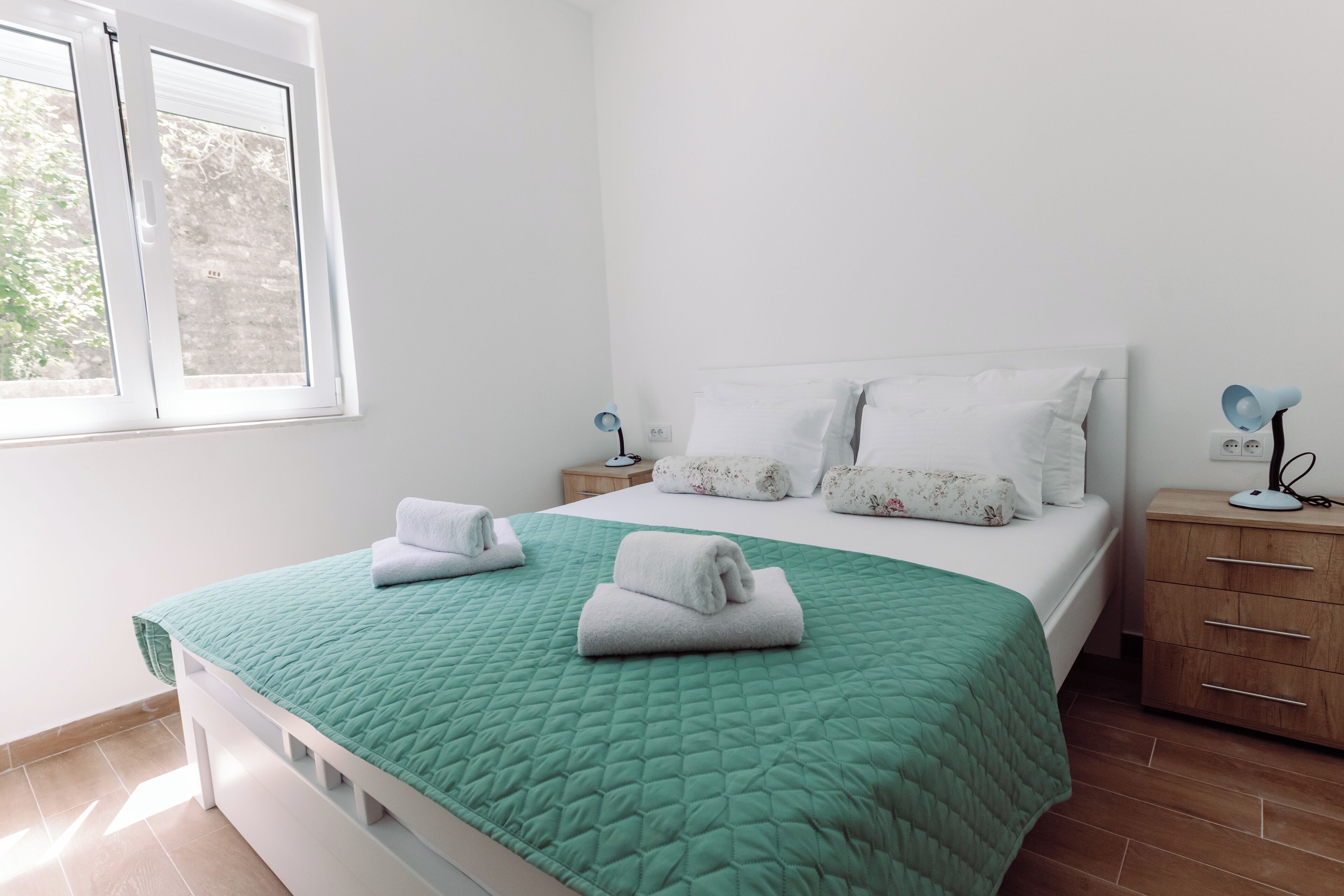 A neatly made bed with a green quilt, white pillows, and two folded towels on the side. There is a bedside table with a lamp and a power outlet.