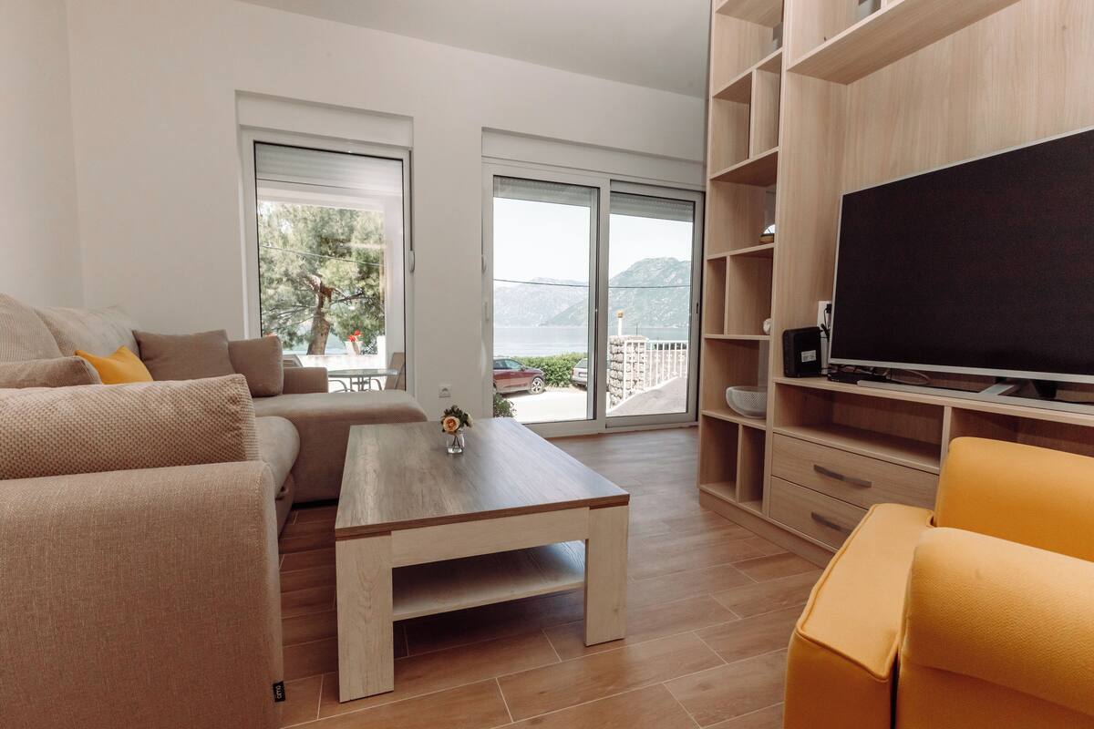 Uma sala de estar moderna com um sofá, mesa de centro e TV. Há portas de vidro deslizantes que dão para uma varanda com vista para as montanhas.