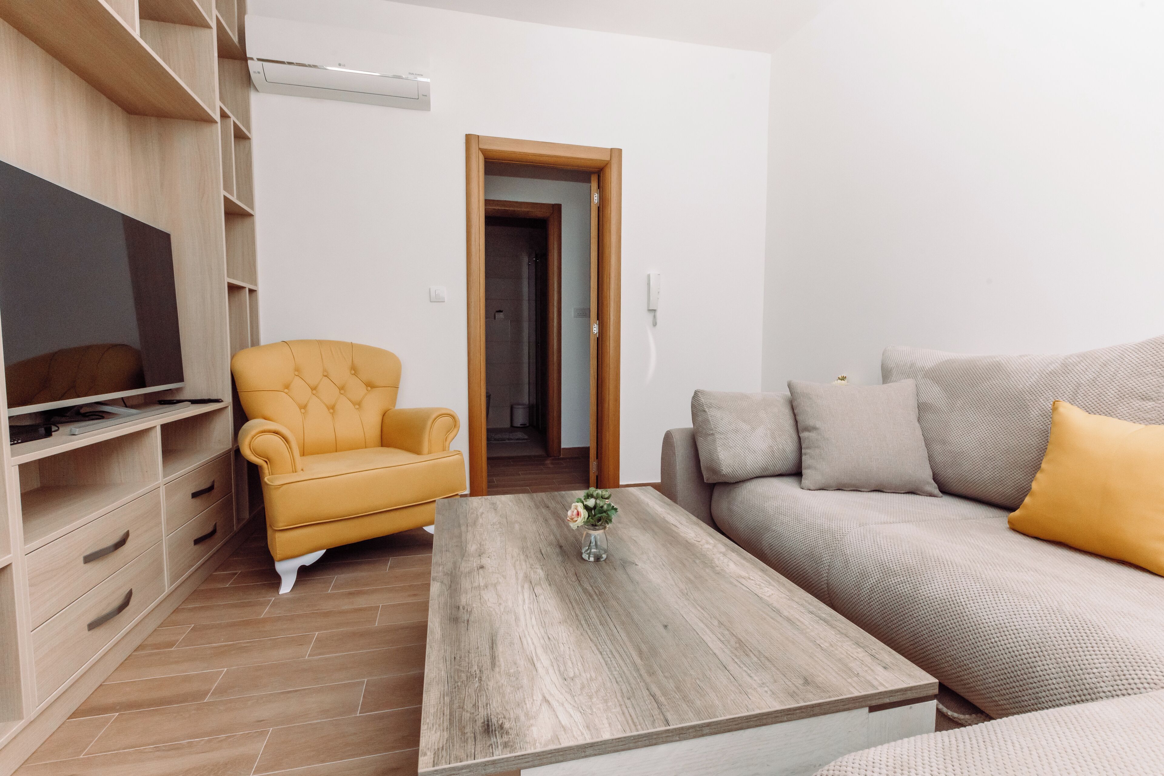 A modern living room with a wooden coffee table, a yellow armchair, a beige sofa with yellow cushions, a television on a wooden shelf, and a wooden wardrobe.