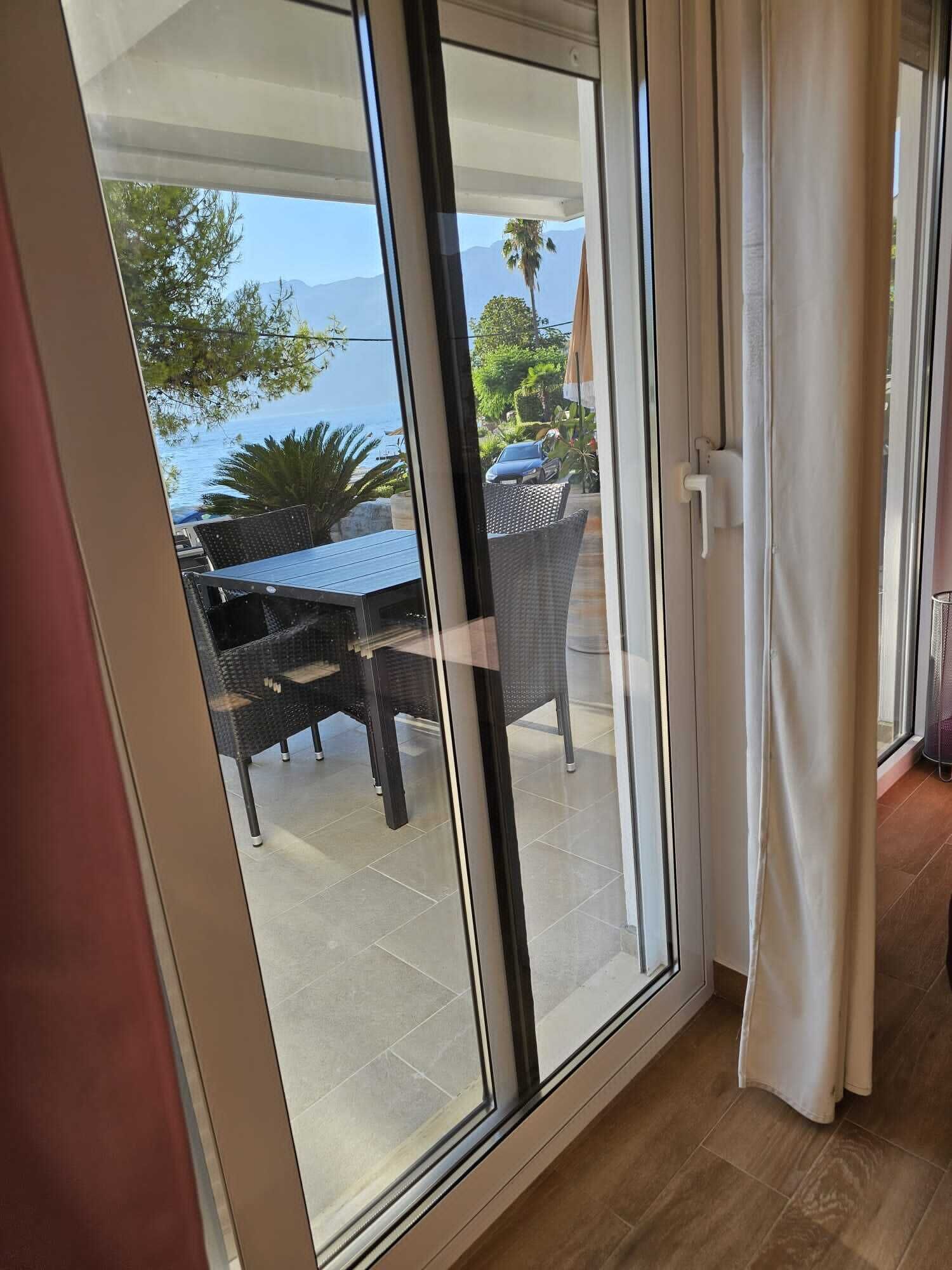 A sliding glass door leading to a patio with a table and chairs, a car, and a view of the sea and mountains.