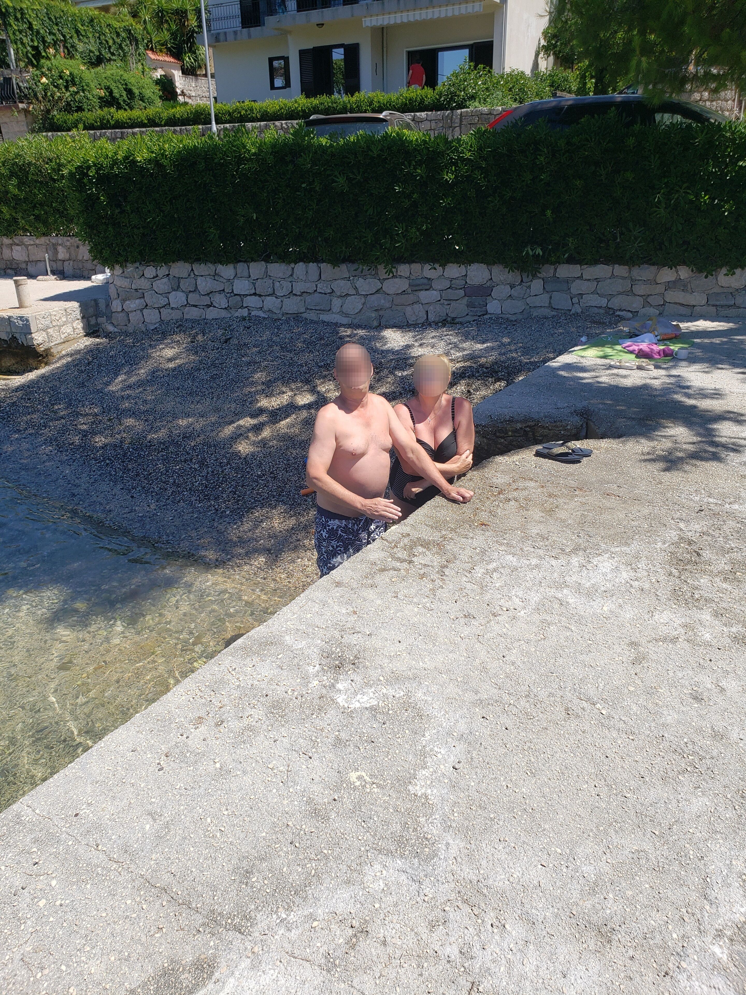 Un hombre y una mujer están junto a una piscina, con un muro de piedra y vegetación al fondo.