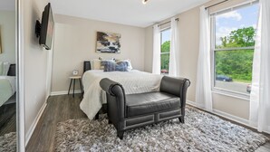 Une chambre à coucher avec un lit, un fauteuil en cuir noir, un tableau, une horloge et une fenêtre donnant sur un paysage verdoyant.