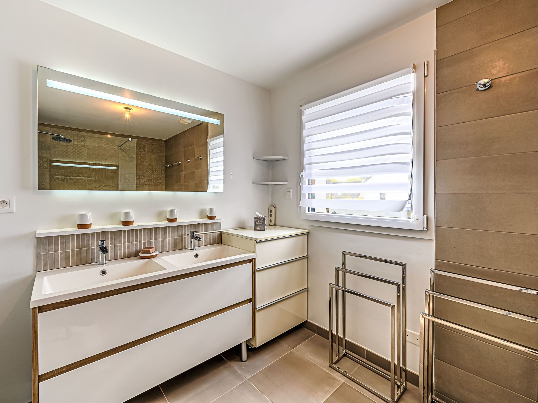A modern bathroom with a large mirror, two sinks, a towel rack, and a window with blinds.