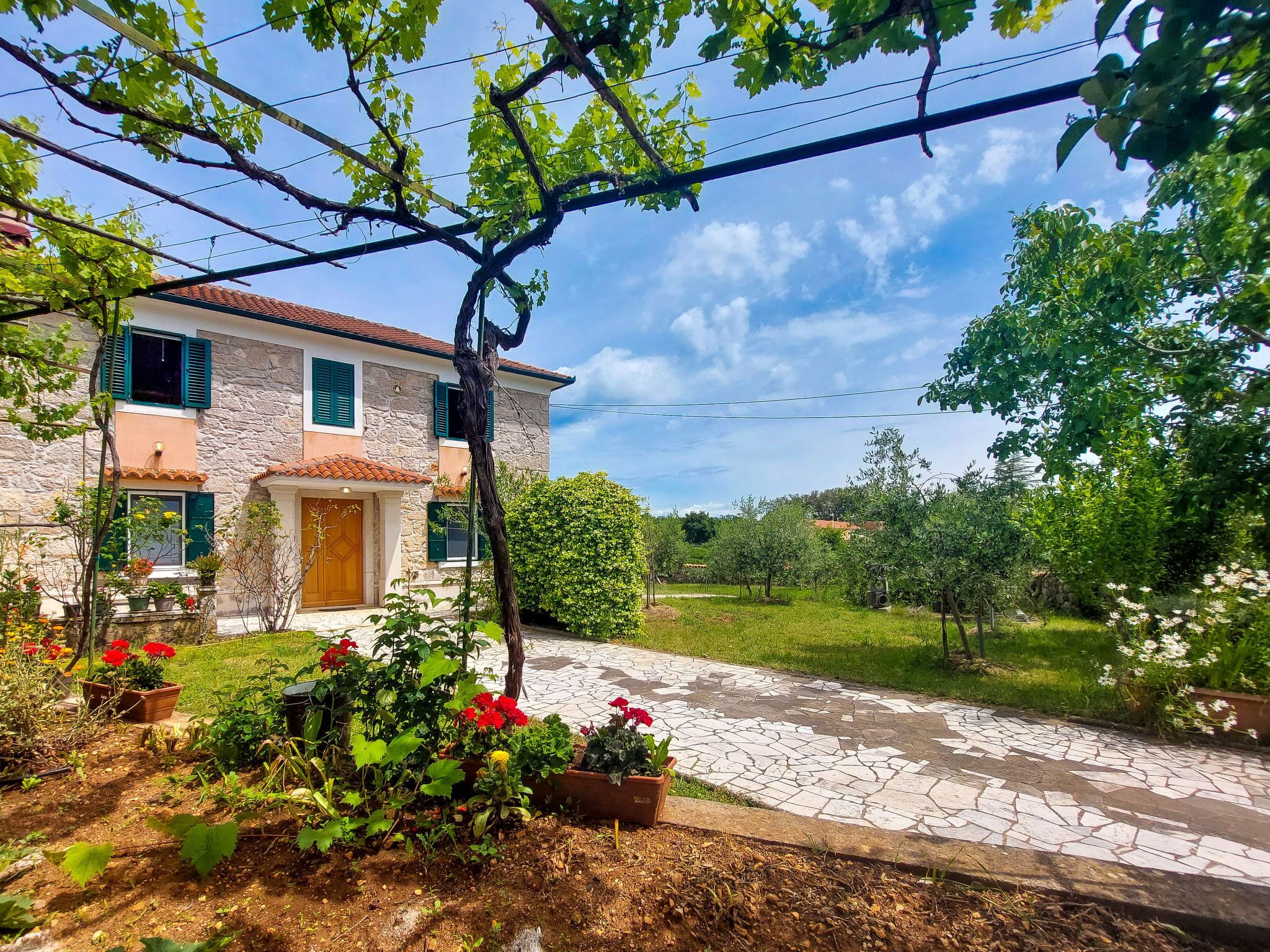 A two-story stone house with a tiled roof, a stone pathway, and a garden with potted plants and flowers.