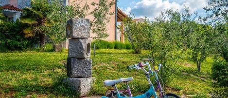 A turquoise bicycle parked in front of a stone pillar, with a house and lush greenery in the background.