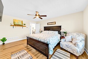 A bedroom with a bed, a chair, a ceiling fan, a painting, and a door leading to another room.
