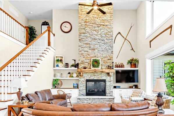 A living room with a stone fireplace, leather sofa, wooden staircase, and a flat-screen TV.