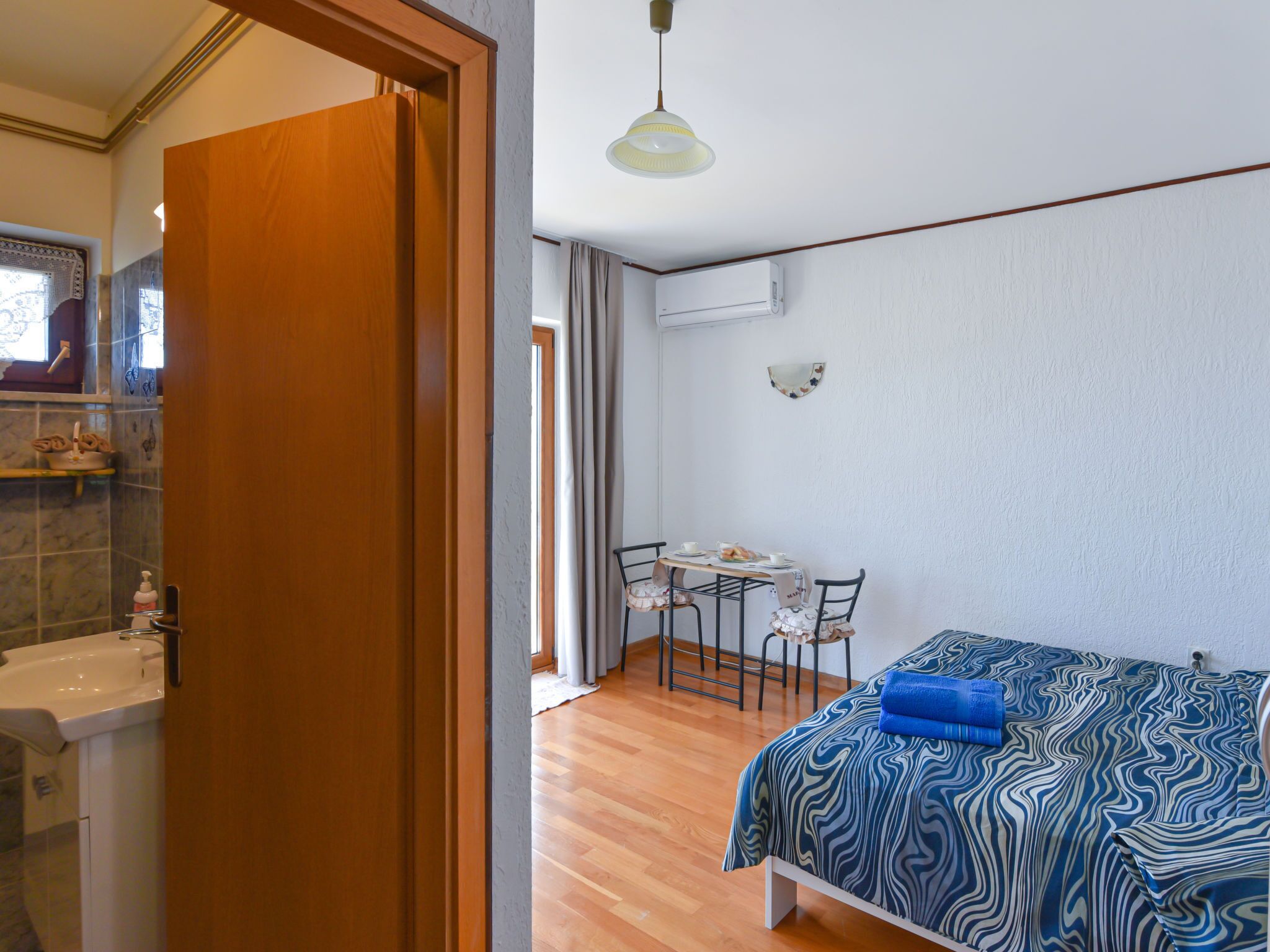 Quarto com cama, mesa de jantar com cadeiras, banheiro com pia e espelho, e corredor que dá para outro cômodo.