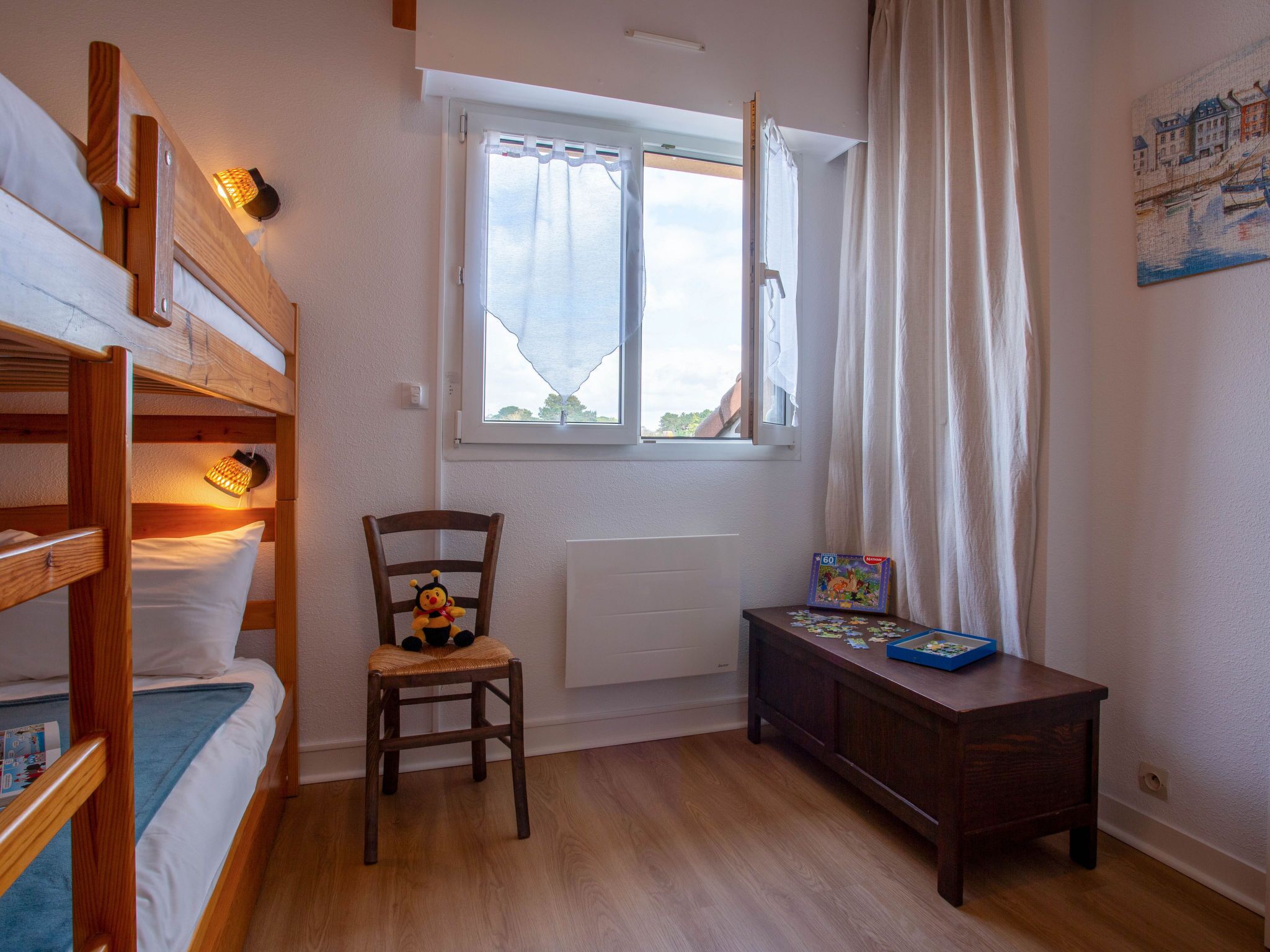 Una habitación con literas, una silla, una mesita con un rompecabezas y una ventana con cortinas.
