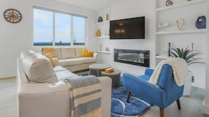 A modern living room with a sofa, armchair, and TV. There is a large window with a view of the sea. - Secord Serenity (Edmonton)