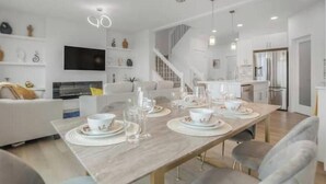 A modern dining area with a long table set for four, a staircase, and a kitchen area with stainless steel appliances. - Secord Serenity (Edmonton)