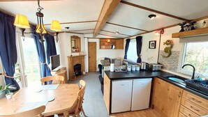 Cocina compacta con armarios de madera, una mesa de comedor con sillas, un refrigerador pequeño y un ventanal con cortinas.