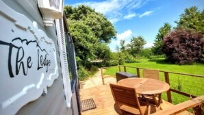 A wooden deck with a table and chairs, a sign reading "The Lodge," and a view of a green lawn and trees.