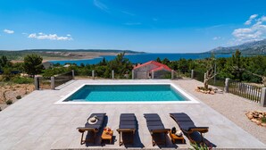 Ein rechteckiger Pool mit Liegestühlen und Sonnenliegen, umgeben von einem Zaun, mit Blick auf Meer und Berge im Hintergrund.