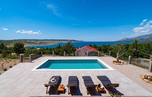 A rectangular swimming pool with lounge chairs and sun loungers, surrounded by a fence, with a view of the sea and mountains in the background. - Nice home in Rovanjska (Rovanjska)