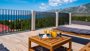 Ein Holztisch mit einem Tablett Orangensaft und einer Flasche, eine hölzerne Bank und Blick auf das Meer und die Berge.