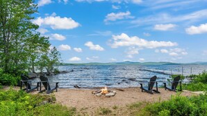 Un cadre idyllique au bord du lac, avec un foyer et des chaises disposées face à l’eau, le tout entouré de verdure et sous un ciel dégagé.