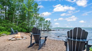 Deux fauteuils Adirondack sur une plage de sable, avec un foyer et une vue sur un lac et une forêt.