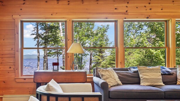 L’intérieur chaleureux d’un chalet, avec un revêtement mural en bois, un canapé, un fauteuil et une table. De grandes fenêtres offrent une vue sur les arbres et un plan d’eau.