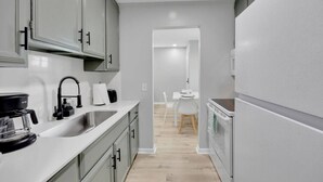 A narrow kitchen with grey cabinets, a sink, a stove, and a dining area in the background.